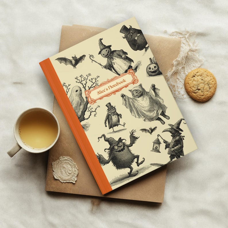 May include: A notebook with an orange spine and a cream cover, illustrated with vintage Halloween characters. The cover features various spooky figures and the text "Alice's Notebook". A cup of tea and a biscuit are also visible.