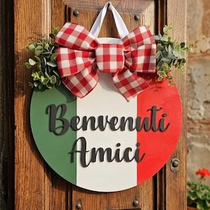 May include: A round wooden welcome sign with the colors of the Italian flag: green, white, and red. The sign has the words "Benvenuti Amici" in black script. A red and white checkered bow and greenery are attached to the top.