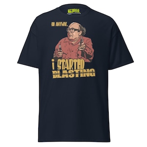 May include: Navy blue t-shirt featuring a graphic of a man holding two guns, with the text "GO ANYWAY." and "I STARTED BLASTING" in yellow. The shirt has a crew neck and short sleeves.