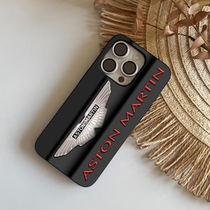 Puede incluir: Funda de teléfono negra con el logotipo de Aston Martin en plata y las palabras "ASTON MARTIN" en rojo. La funda tiene un diseño en relieve y está hecha de un material duradero. La funda está diseñada para proteger el teléfono de arañazos y daños.