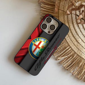 Puede incluir: Funda de teléfono negra con el logotipo de Alfa Romeo, un diseño circular con detalles rojos y blancos. La funda tiene la inscripción "Alfa Romeo Racing" en el lateral.