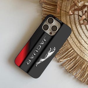 Puede incluir: Funda de teléfono negra con el logotipo de Jaguar. La funda tiene una sección negra texturizada, un degradado rojo y el logotipo de Jaguar en plata. Las lentes de la cámara del teléfono son visibles en la parte superior.
