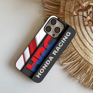 Puede incluir: Funda de teléfono negra con el logotipo de Honda Racing en rojo, blanco y azul. La funda tiene un acabado brillante y un borde negro. La funda del teléfono está sobre una superficie tejida.