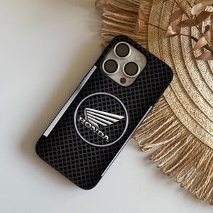 May include: Black iPhone case with a textured diamond pattern and a silver Honda logo. The logo features the Honda name and a stylized wing emblem within a silver circle. The case has a silver stripe along the side.