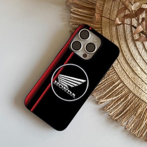 Może przedstawiać: Czarne etui na telefon z czerwonym paskiem i logo Hondy. Logo to srebrna odznaka ze słowem "HONDA" pod wzorem skrzydła. Etui ma błyszczące wykończenie i jest przeznaczone do smartfona.