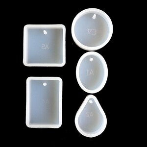 Op de afbeelding: Set van vier witte siliconen mallen voor het maken van sieraden hangers. De mallen zijn gevormd als een cirkel, ovaal, rechthoek en traan. Elke mal heeft een klein gaatje bovenaan om de hanger op te hangen.