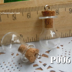 Può includere: Due piccole bottiglie di vetro con tappi di sughero e tappi argentati. Le bottiglie sono alte circa 3,8 cm e sono appoggiate su una superficie di pizzo bianco. Le bottiglie sono etichettate con "P006".