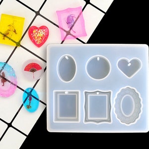 Puede incluir: Un molde de silicona transparente con siete formas diferentes para hacer joyas de resina. Las formas incluyen un corazón, un óvalo, un círculo, un cuadrado, un rectángulo y dos marcos diferentes. El molde está sobre una superficie de azulejos blancos.