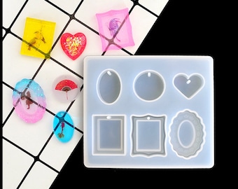 Puede incluir: Un molde de silicona transparente con siete formas diferentes para hacer joyas de resina. Las formas incluyen un corazón, un óvalo, un círculo, un cuadrado, un rectángulo y dos marcos diferentes. El molde está sobre una superficie de azulejos blancos.