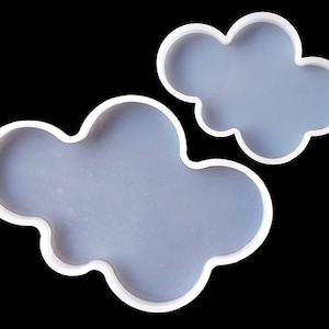Puede incluir: Dos moldes de silicona blanca con forma de nubes. El molde de nube más grande está en la parte inferior y el molde de nube más pequeño está en la parte superior.