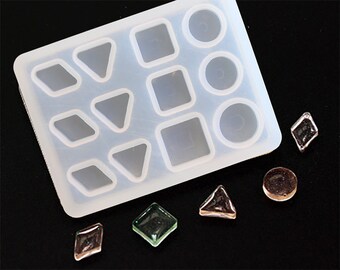 May include: A white silicone mold with 12 cavities for making resin jewelry. The cavities are shaped like circles, squares, and triangles. There are also five resin pieces in different shapes and colors next to the mold.