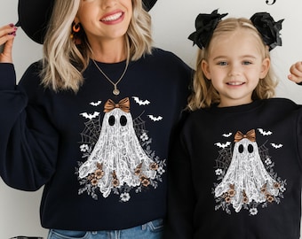 May include: Black sweatshirts with a ghost design. The ghost is white with a brown bow, surrounded by flowers, bats, and spiderwebs. The word "BLACK" is printed at the bottom of the image.