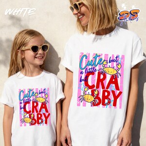 May include: White t-shirts with the text "Cute but a little crabby" in blue and red, featuring yellow crab illustrations. The design is set against a pink striped background.