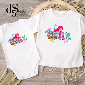 Puede incluir: Un body blanco de manga corta y una camiseta blanca de manga corta, ambos con un gr&aacute;fico colorido de una sirena con cabello rosa y el nombre EMILY en letras multicolores estampadas, decorado con corazones, estrellas y flores.