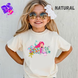 Könnte beinhalten: Cremefarbenes T-Shirt mit einem bunten Meerjungfrau-Motiv und dem Namen "Emily". Das Design umfasst Herzen, Blumen und eine Schleife. Das Wort "NATURAL" ist oben rechts in Schwarz gedruckt.