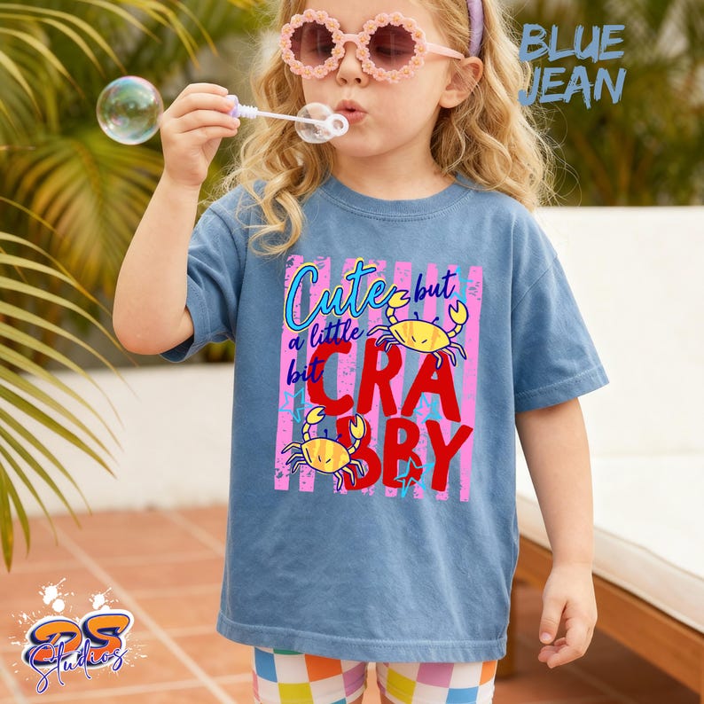 May include: A blue jean t-shirt with the text "Cute but a little bit Crabby" and two yellow crab illustrations. The shirt is worn by a child blowing bubbles and wearing pink sunglasses.