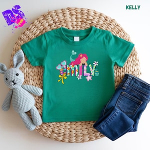 Puede incluir: Una camiseta de manga corta de color verde esmeralda con un gr&aacute;fico de una sirena de pelo rosa y el nombre EMILY en letras coloridas con patrones. El dise&ntilde;o incluye peque&ntilde;as flores, corazones y un lazo amarillo. La camiseta est&aacute; sobre una estera tejida.