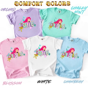 Puede incluir: Cinco camisetas de manga corta en colores orqu&iacute;dea, menta tiza, flor, blanco y chambray. Cada camiseta presenta un gr&aacute;fico de una sirena con cabello rosa sobre el nombre Jane u Olivia en letras coloridas, rodeado de corazones y flores.