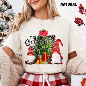 Może przedstawiać: Kremowa koszulka z bożonarodzeniowym wzorem. Grafika zawiera choinkę, dwa gnomy, laski cukrowe i zapakowane prezenty. Tekst "TIS THE SEASON" jest wydrukowany nad drzewem czarnym pismem.