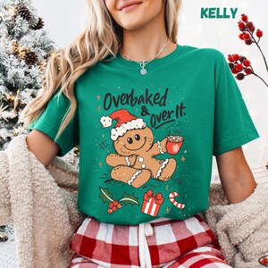May include: A green t-shirt with a gingerbread man graphic wearing a Santa hat and holding a mug. The text "Overbaked & Over It" is above the gingerbread man. Christmas-themed illustrations, including holly, a gift, and a candy cane, are also featured.