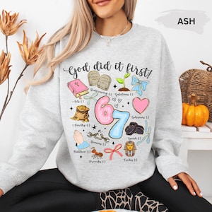 Könnte beinhalten: Hellgraues Sweatshirt mit dem Schriftzug "God did it first!" und verschiedenen religiösen Illustrationen. Das Design umfasst ein rosa Herz, eine blaue Schleife und die Zahlen "67". Ein bequemes Sweatshirt im lässigen Stil.