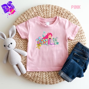Puede incluir: Una camiseta de manga corta de color rosa claro con un gr&aacute;fico de una sirena de pelo rosa y el nombre Emily en letras de bloque coloridas y estampadas. El dise&ntilde;o incluye peque&ntilde;os elementos decorativos como corazones, flores y un lazo amarillo.