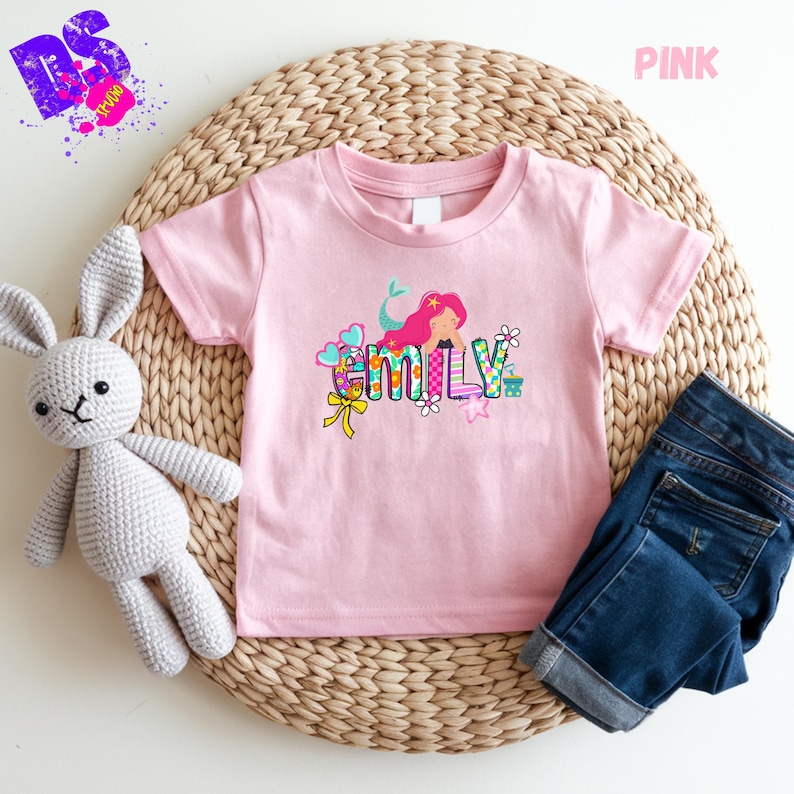 Puede incluir: Una camiseta de manga corta de color rosa claro con un gr&aacute;fico de una sirena de pelo rosa y el nombre Emily en letras de bloque coloridas y estampadas. El dise&ntilde;o incluye peque&ntilde;os elementos decorativos como corazones, flores y un lazo amarillo.