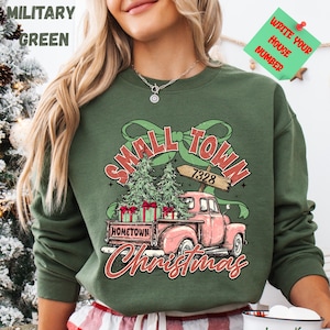 May include: Military green sweatshirt with a graphic featuring a pink vintage truck carrying Christmas trees and gifts. The text reads "Small Town Christmas" and "Hometown". A green sticky note says "Write Your House Number".