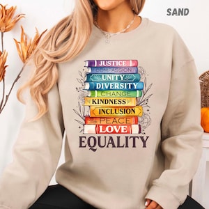 May include: Sand-colored sweatshirt featuring a stack of books with words like "Justice," "Equality," and "Love." The books are in various colors and are decorated with floral accents. The word "EQUALITY" is printed in large letters at the bottom.