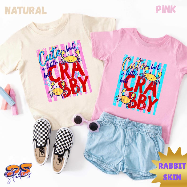 May include: Two t-shirts, one natural and one pink, with the text "Cute but a little Crabby" and crab graphics. The image also includes denim shorts, checkered slip-on shoes, and sunglasses.