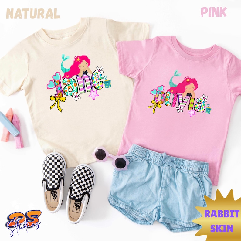 Puede incluir: Dos camisetas de manga corta, una natural y otra rosa, cada una con un gr&aacute;fico de una sirena y los nombres Jane u Olivia. Las camisetas van acompa&ntilde;adas de pantalones cortos de mezclilla azul claro, zapatos sin cordones a cuadros y gafas.