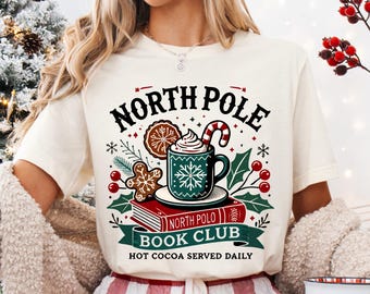 Puede incluir: Camiseta color crema con un diseño festivo. El diseño presenta las palabras "North Pole Book Club" con una taza de chocolate caliente, galletas de jengibre y bastones de caramelo. También incluye el texto "Hot Cocoa Served Daily".