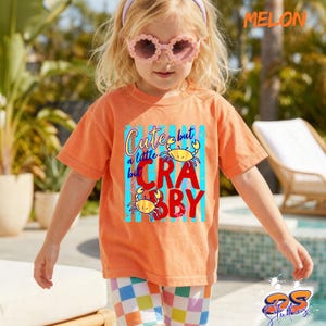 May include: An orange t-shirt with the text "Cute but a little crabby" and two cartoon crabs. The shirt is worn by a child wearing pink sunglasses and a purple headband. The child is also wearing checkered shorts.