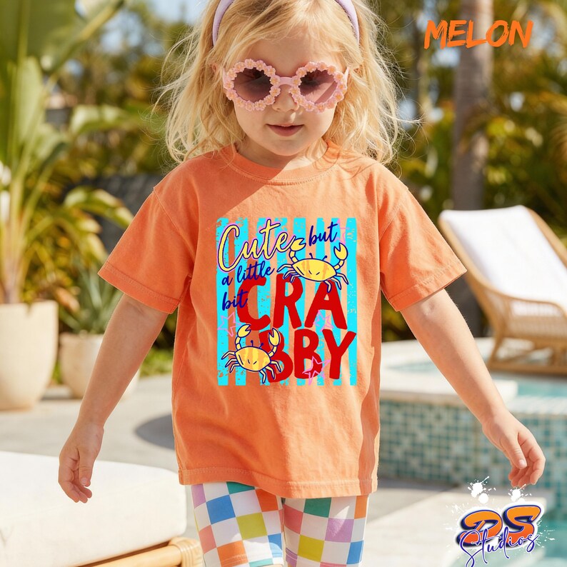 May include: An orange t-shirt with the text "Cute but a little crabby" and two cartoon crabs. The shirt is worn by a child wearing pink sunglasses and a purple headband. The child is also wearing checkered shorts.