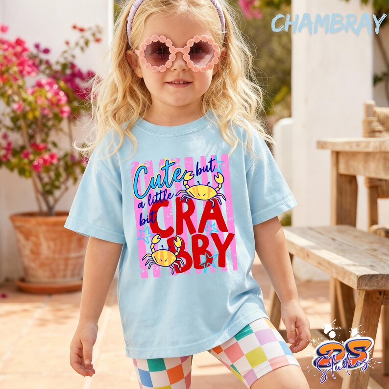 May include: A young girl wearing a light blue t-shirt with the text "Cute but a little Crabby" and crab graphics. She is also wearing pink sunglasses and colorful shorts. The word "Chambray" is printed in the upper right corner.