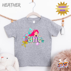 Puede incluir: Una camiseta gris jaspeado de manga corta con un estampado gr&aacute;fico colorido de una sirena con cabello rosa brillante. El nombre Jane est&aacute; escrito en letras grandes y estampadas decoradas con flores, corazones y un lazo amarillo. Texto Rabbit Skin.