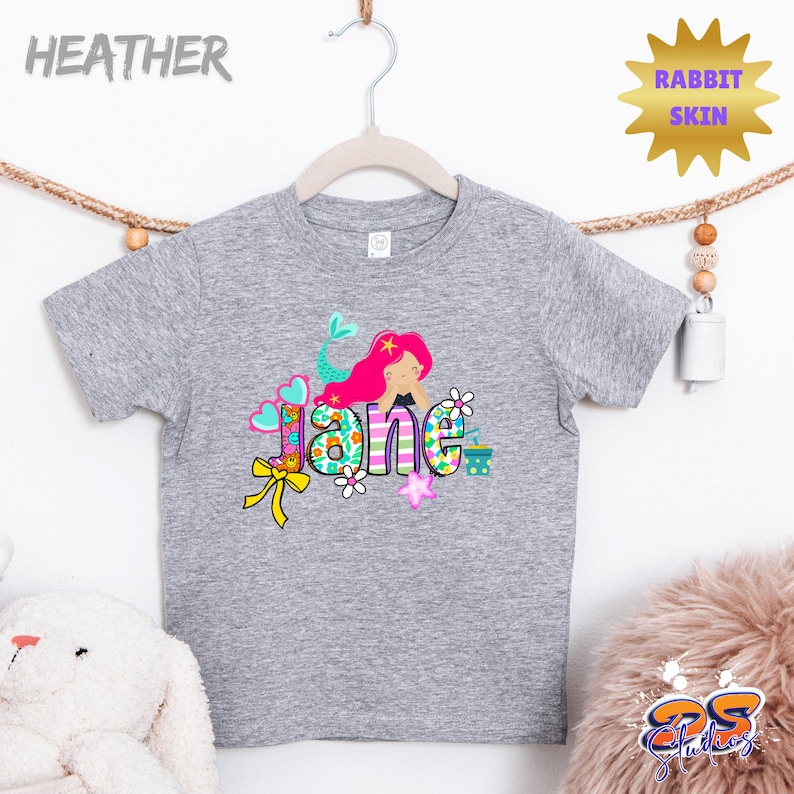 Puede incluir: Una camiseta gris jaspeado de manga corta con un estampado gr&aacute;fico colorido de una sirena con cabello rosa brillante. El nombre Jane est&aacute; escrito en letras grandes y estampadas decoradas con flores, corazones y un lazo amarillo. Texto Rabbit Skin.