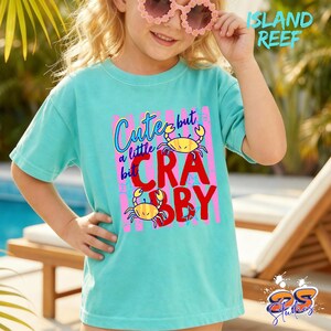 May include: A turquoise t-shirt with the text "Cute but a little Crabby" in a colorful design featuring two yellow crabs. The shirt is worn by a person wearing pink sunglasses. The words "Island Reef" are in the upper right corner.
