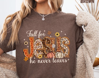 May include: Heather brown t-shirt featuring the text "Fall for Jesus he never leaves." The word "Jesus" is designed with a leopard print pattern, complemented by autumnal elements like pumpkins and sunflowers. A fall-themed religious garment.