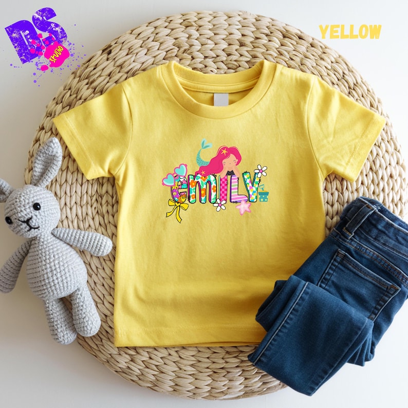 Puede incluir: Una camiseta amarilla de manga corta con un estampado gr&aacute;fico colorido del nombre Emily en letras estilizadas, decorada con una sirena de pelo rosa, peque&ntilde;as flores, corazones y un lazo amarillo, acompa&ntilde;ada de unos vaqueros azul oscuro.