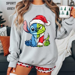 May include: Sport grey sweatshirt featuring a split cartoon character design. One side is blue, the other green, both wearing a Santa hat and coat. The sweatshirt has the text "SPORT GREY" at the top. The person is holding a mug with Christmas trees.