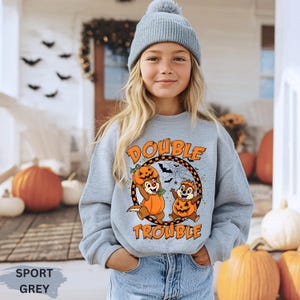 May include: A light grey sweatshirt with the text "DOUBLE TROUBLE" and cartoon characters dressed as pumpkins. The sweatshirt is worn by a person wearing a grey beanie. The word "SPORT GREY" is printed at the bottom.
