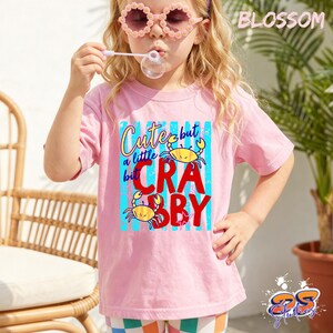 May include: A pink t-shirt with the text "Cute but a little crabby" and crab graphics. The shirt is worn by a child wearing pink flower-shaped sunglasses and blowing bubbles. The word "BLOSSOM" is printed at the top right.