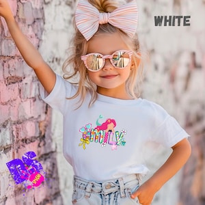 Puede incluir: Una camiseta blanca de manga corta con un estampado gr&aacute;fico colorido del nombre EMILY en letras de bloque, decorado con una sirena de pelo rosa, flores, corazones y un lazo amarillo. La camiseta se combina con unos vaqueros azul claro.
