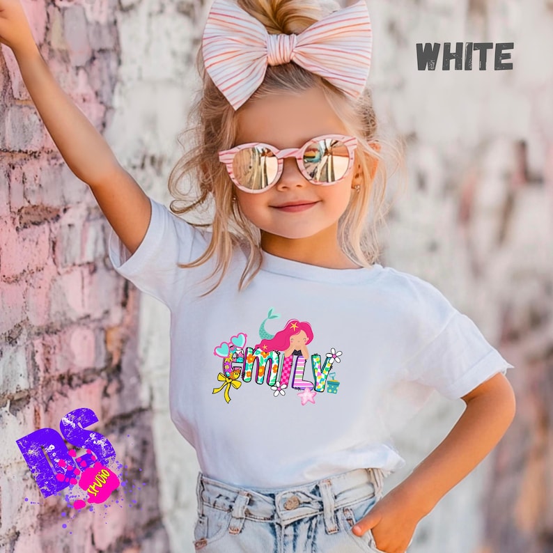 Puede incluir: Una camiseta blanca de manga corta con un estampado gr&aacute;fico colorido del nombre EMILY en letras de bloque, decorado con una sirena de pelo rosa, flores, corazones y un lazo amarillo. La camiseta se combina con unos vaqueros azul claro.