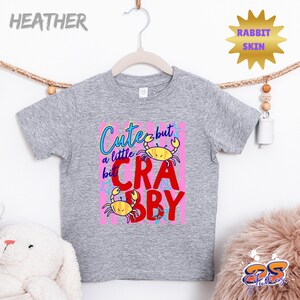 May include: A heather gray toddler t-shirt with a colorful graphic. The graphic reads "Cute but a little Crabby" with yellow crab illustrations on a pink background. The shirt is hanging on a wooden hanger.