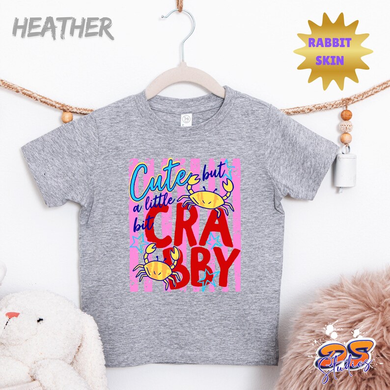 May include: A heather gray toddler t-shirt with a colorful graphic. The graphic reads "Cute but a little Crabby" with yellow crab illustrations on a pink background. The shirt is hanging on a wooden hanger.