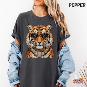 Könnte beinhalten: Ein anthrazitfarbenes T-Shirt mit einem Grafikdruck eines Tigers, der eine Brille mit Leopardenmuster und eine große Halskette im Kristallstil trägt. Das Wort PEPPER steht in der oberen rechten Ecke. Das T-Shirt wird mit einer hellblauen Jeansjacke getragen.
