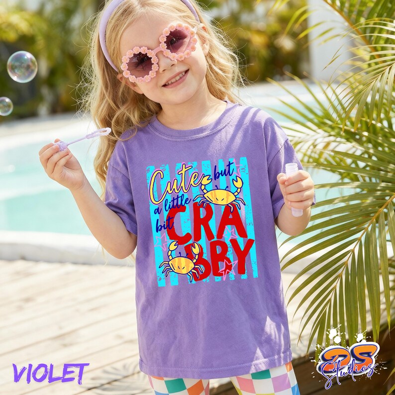 May include: A violet t-shirt with the text "Cute but a little Crabby" in a colorful design. The shirt features two yellow crab illustrations. The image also shows a young person wearing the shirt and sunglasses.