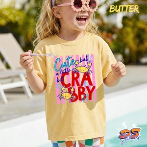 May include: A yellow t-shirt with the text "Cute but a little Crabby" in a colorful design featuring crabs. The shirt is worn by a child wearing pink sunglasses and a headband. The word "Butter" is printed in the upper right corner.